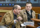 Todd Johnson sits at the testifiers' table and speaks to someone out of view while gesturing with a hand. Representative Lillie sits next to him and watches as he speaks.